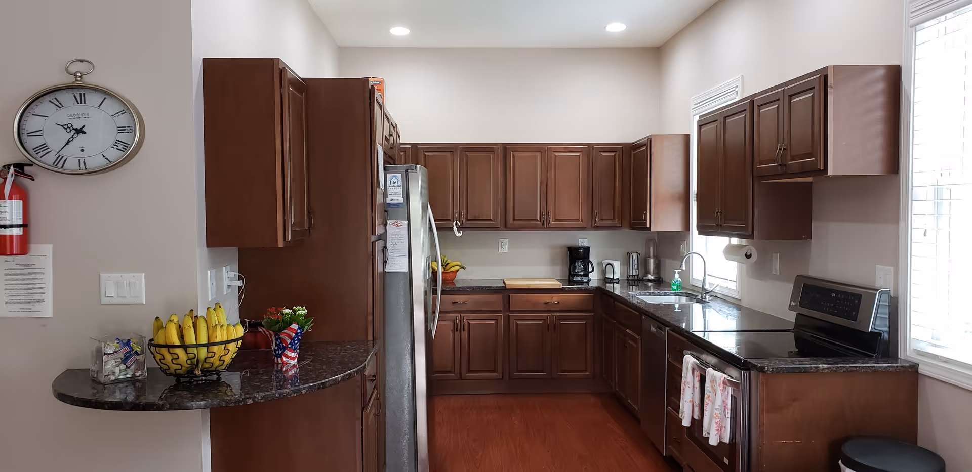 A modern kitchen with dark wooden cabinets, a stainless steel refrigerator, a stove with two towels hanging on the handle, a coffee maker, and a sink under a window with blinds. There is a wall clock, a fire extinguisher, a bowl of bananas, and a small potted plant on a curved countertop.