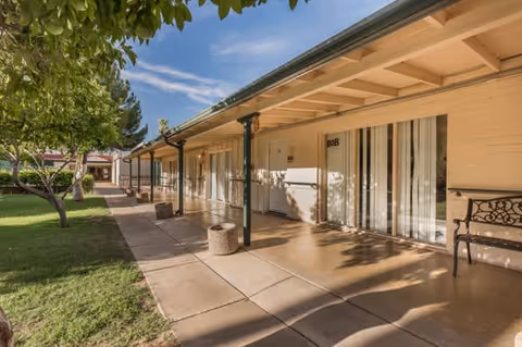 Outdoor walkway at Maryland Gardens Assisted Living with a covered patio supported by green columns, doors to individual rooms including one labeled 808, benches, and a grassy area with trees under a blue sky.