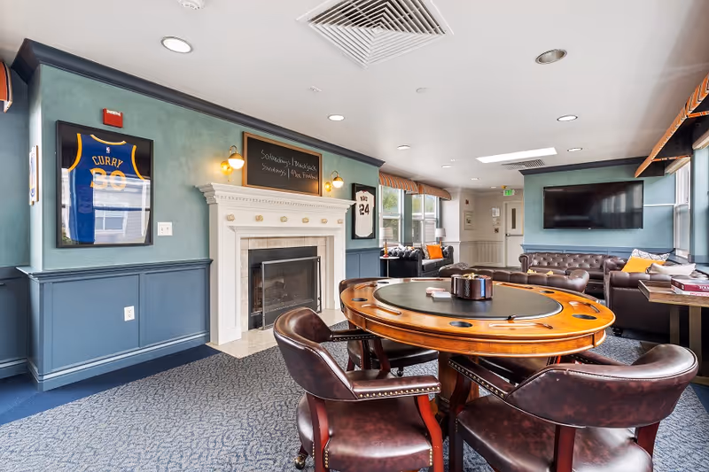 A cozy common area with a round wooden game table surrounded by four leather chairs, a white fireplace with a chalkboard above it, framed sports jerseys on the walls, a large flat-screen TV mounted on the wall, and leather sofas near windows with striped valances.