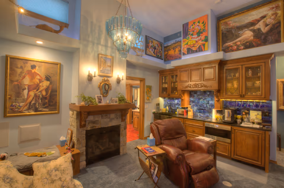 Cozy living area with a leather recliner chair, a stone fireplace with a wooden mantle decorated with plants and framed photos, and a wooden kitchenette with cabinets, a coffee maker, and a blue tiled backsplash. The walls are adorned with various colorful paintings and there is a blue chandelier hanging from the ceiling.
