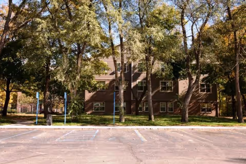 Exterior view of a multi-story brick building partially obscured by tall trees with green and yellow leaves, with an empty parking lot in the foreground including marked handicap parking spaces.