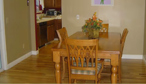 Wooden dining table and chairs in a homey dining area, with a kitchen visible.