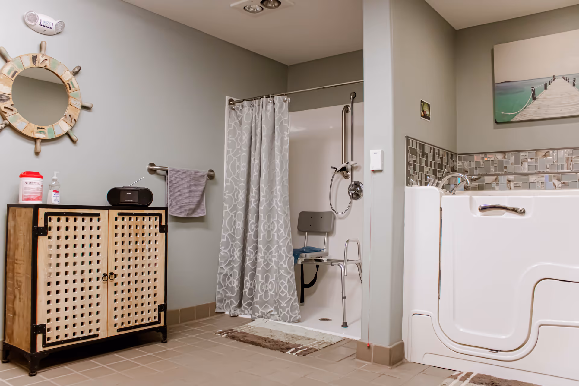 A bathroom designed for assisted living with a walk-in bathtub featuring a door and grab bars, a shower area with a shower chair and a patterned shower curtain, a wooden cabinet with a radio and hygiene products on top, a towel hanging on a wall-mounted rack, a round nautical-themed mirror, and a wall-mounted picture above the bathtub.