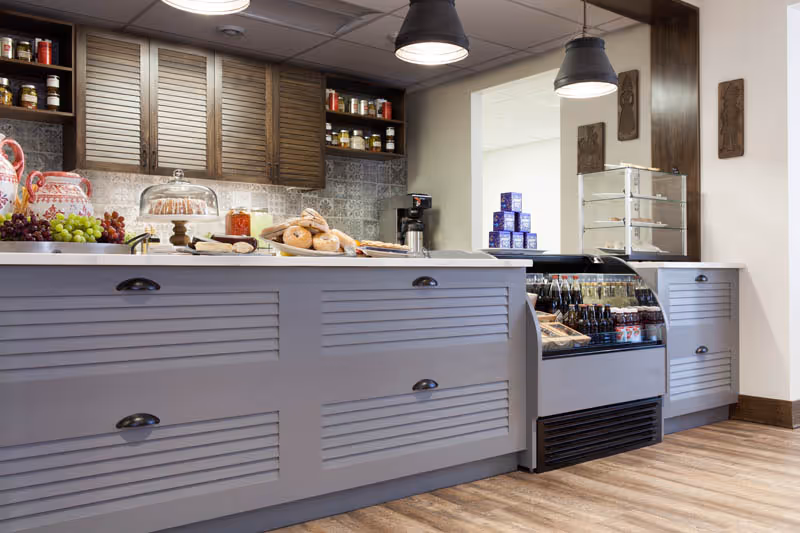 A cafe-style service counter with a display of bagels, fruit, bottled drinks and pendant lights in a dining area.