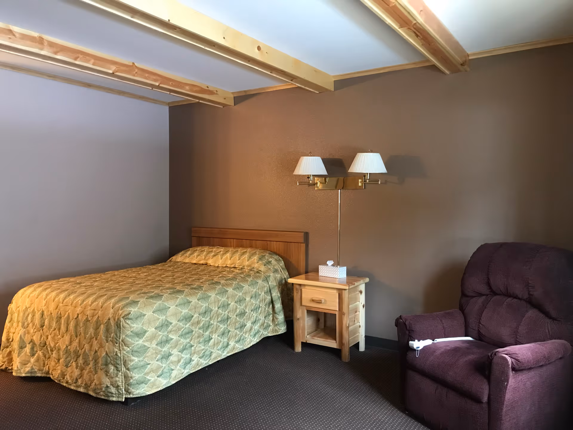 A simple bedroom with a double bed, wooden nightstand and wall-mounted lamps, and a purple recliner under exposed ceiling beams.