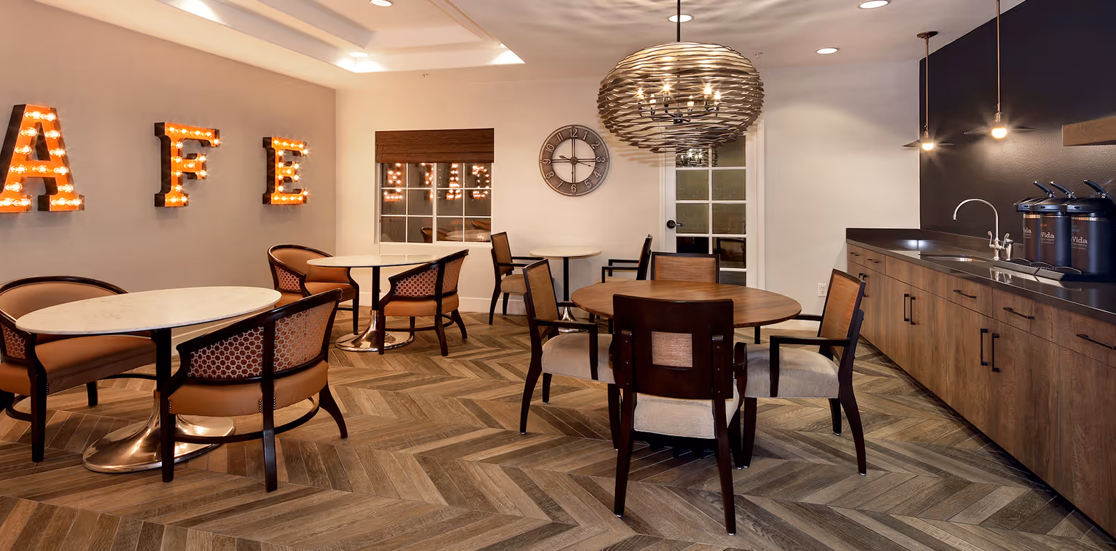 Interior view of a cozy cafe area with round tables and cushioned chairs arranged on a chevron-patterned wooden floor. The wall features illuminated marquee letters spelling 'CAFE'. A large decorative clock hangs on the wall, and a modern pendant light fixture is suspended from the ceiling. To the right, there is a countertop with a sink and beverage dispensers.