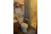 Small bathroom with a toilet, a shower with a curtain, a grab bar, and a shelf above the toilet holding folded towels and other items.