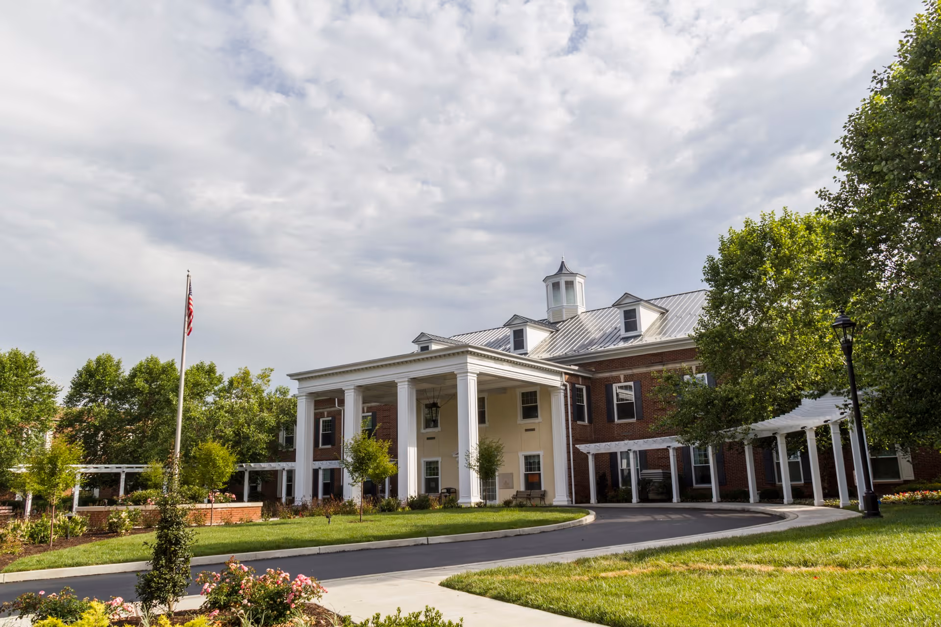 Front exterior view of a large brick building with white columns and a covered entrance, surrounded by green trees, a manicured lawn, and a curved driveway under a cloudy sky.