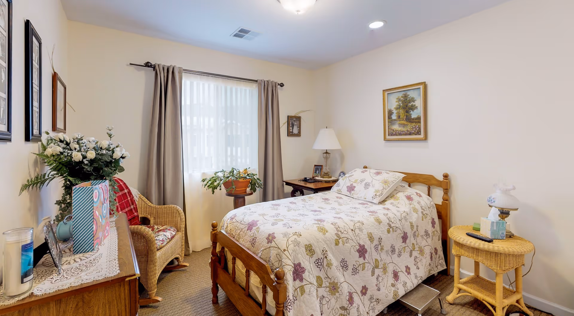 A cozy bedroom with a single wooden bed covered with a floral patterned quilt and matching pillow. There is a wicker chair with a red plaid cushion next to a window with beige curtains. A wooden dresser with a lace runner, flowers, and framed photos is on the left side. A small wicker side table with a lamp, tissues, and a remote control is on the right side. The walls are light-colored with framed artwork hanging.