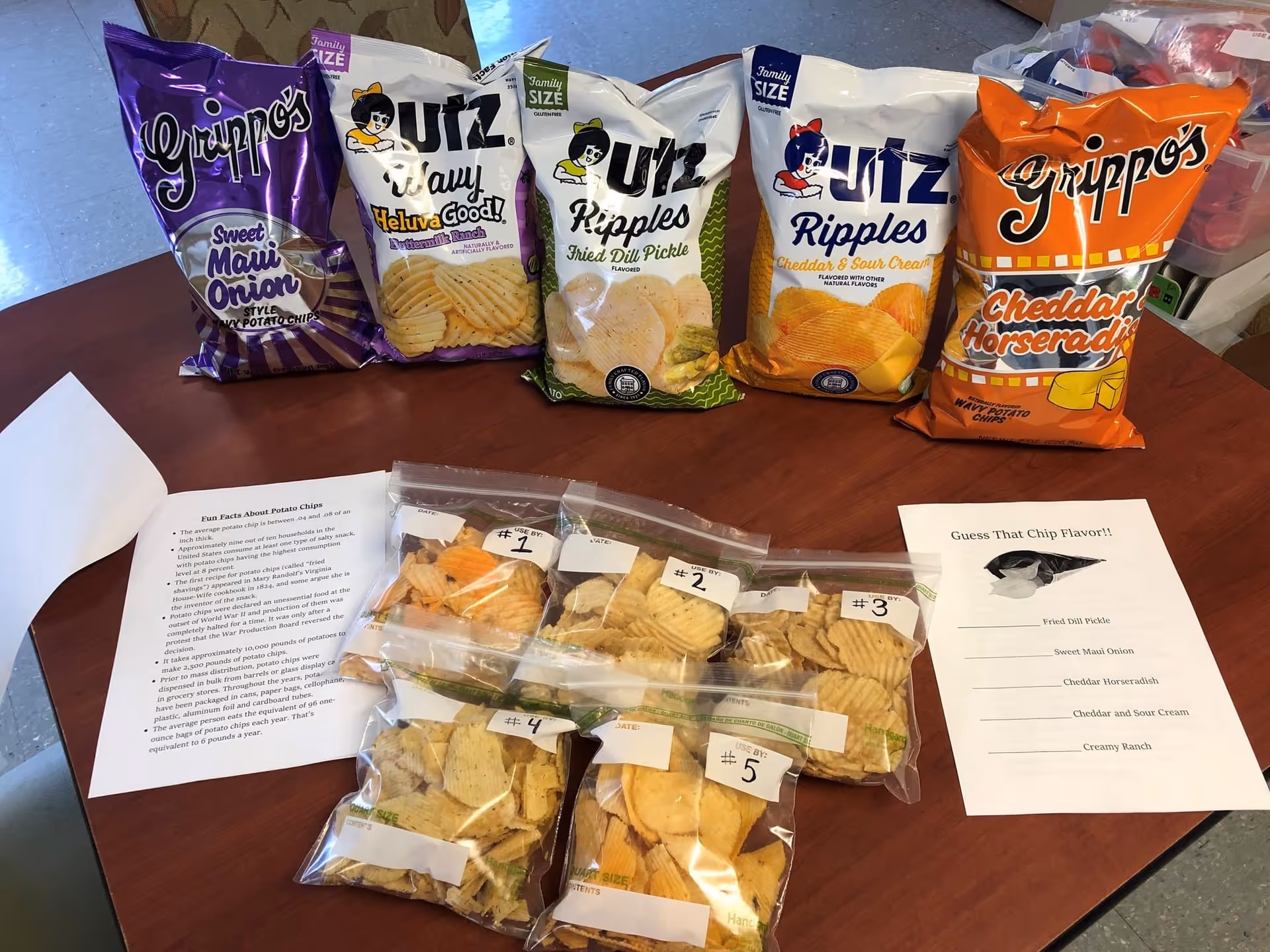 Tabletop chip-tasting setup with five large chip bags, six numbered sample bags in plastic, and printed tasting sheets.