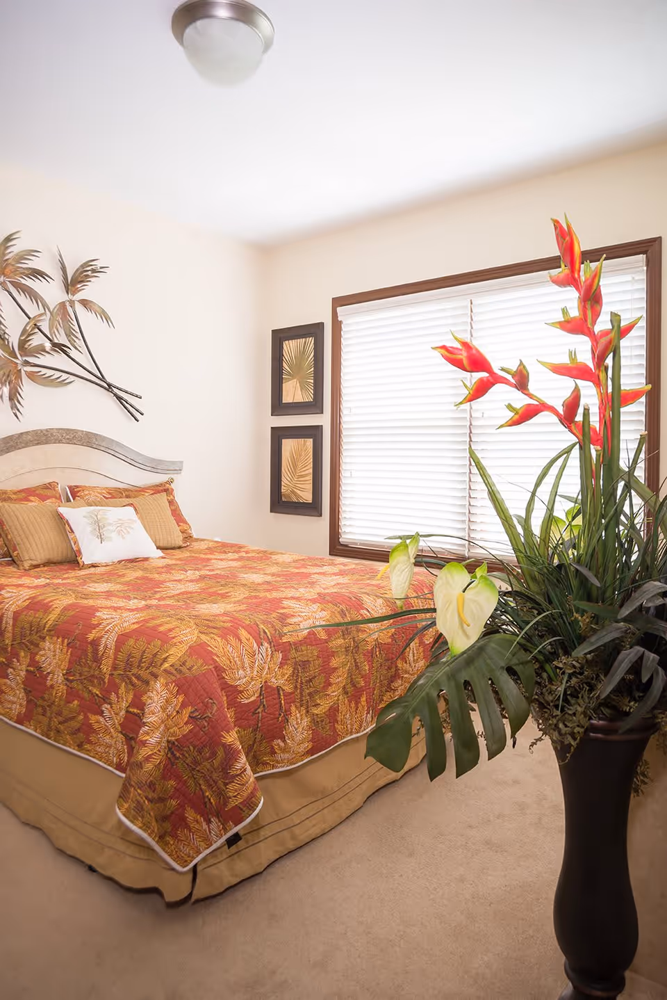 A bedroom with a bed covered in a red and gold leaf-patterned bedspread, matching pillows, and a beige bed skirt. The room has beige walls with two framed leaf prints and a metal wall decoration of palm leaves. A large window with white blinds is on one wall, and a tall black vase with tropical flowers and large green leaves is in the foreground.