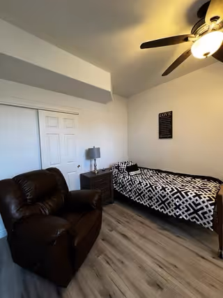 A small bedroom with a single bed covered in a black and white geometric patterned blanket, a dark brown recliner chair, a nightstand with a lamp, and a ceiling fan with a light. The walls are white, and there is a framed inspirational quote on the wall above the bed.