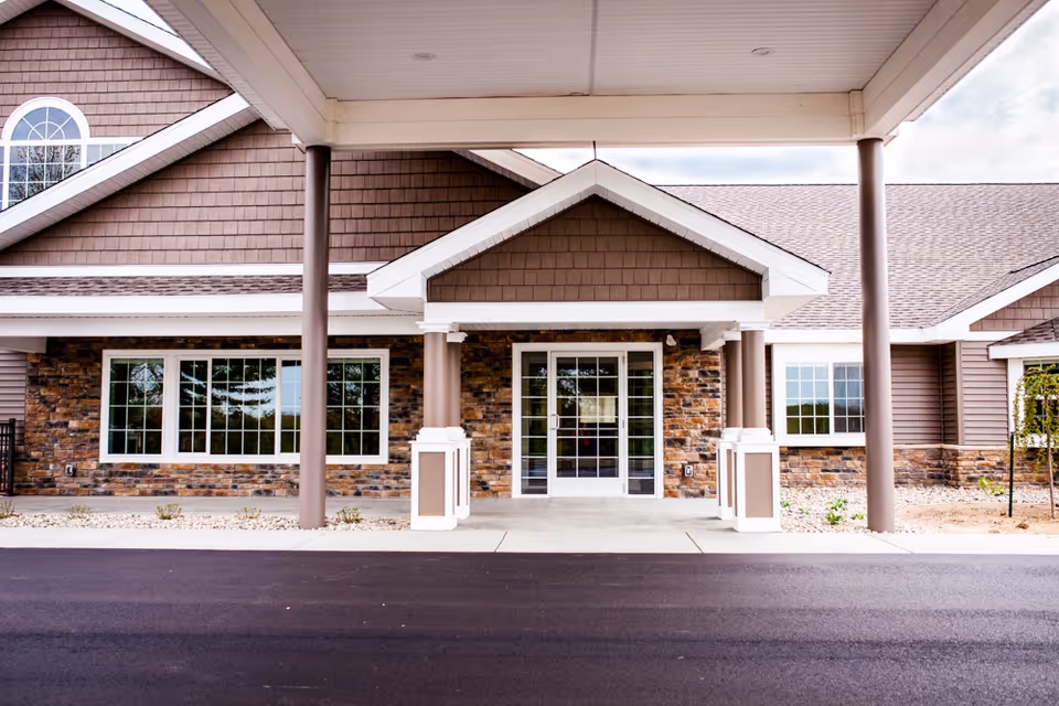 Front exterior entrance of a building with a covered porch supported by columns, large windows, and a double glass door. The building features brown siding and stone accents.