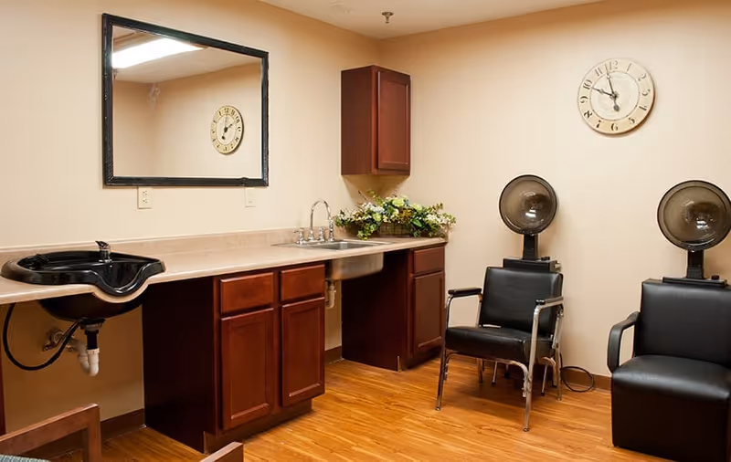 Interior room with wooden flooring featuring a countertop with a sink and dark wood cabinets. Two black salon chairs with hair dryer hoods are positioned against the wall. A large mirror hangs above the countertop and a round wall clock shows the time as 12:05. A floral arrangement is placed on the countertop.