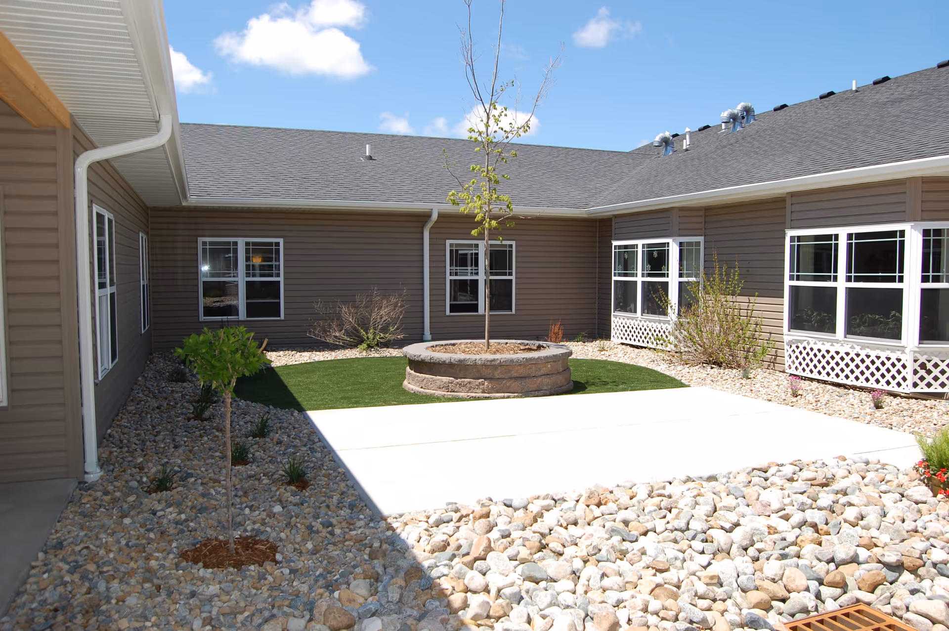 Sunny enclosed courtyard with a central round planter and young tree, rock landscaping, and surrounding building windows.