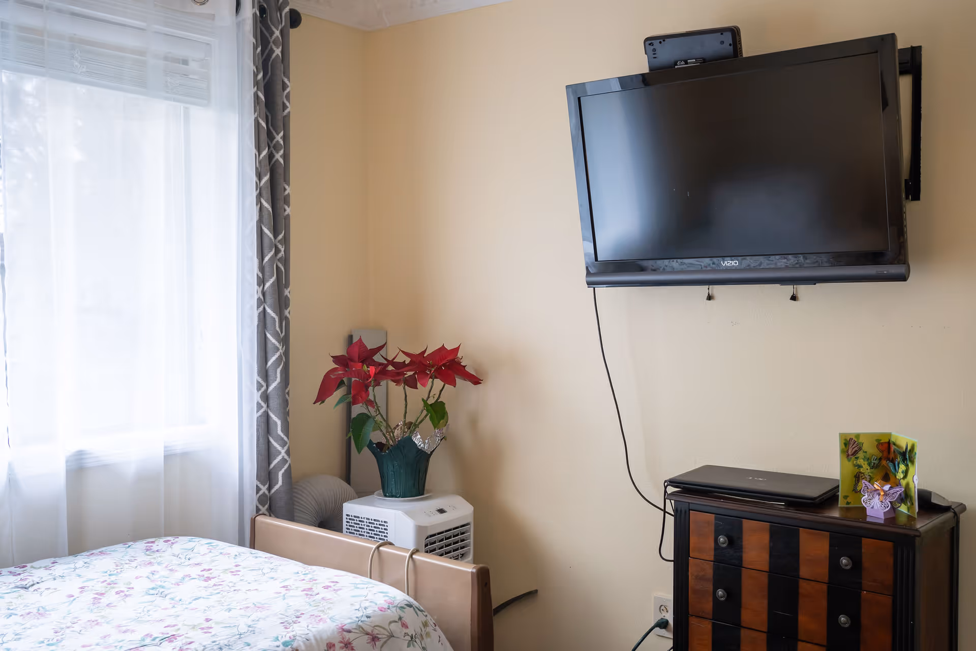 A bedroom with a bed covered in floral bedding near a window with sheer white curtains and gray patterned drapes. A small wooden dresser with a laptop and decorative items sits below a wall-mounted flat-screen TV. A potted poinsettia plant is placed on a white appliance next to the bed.