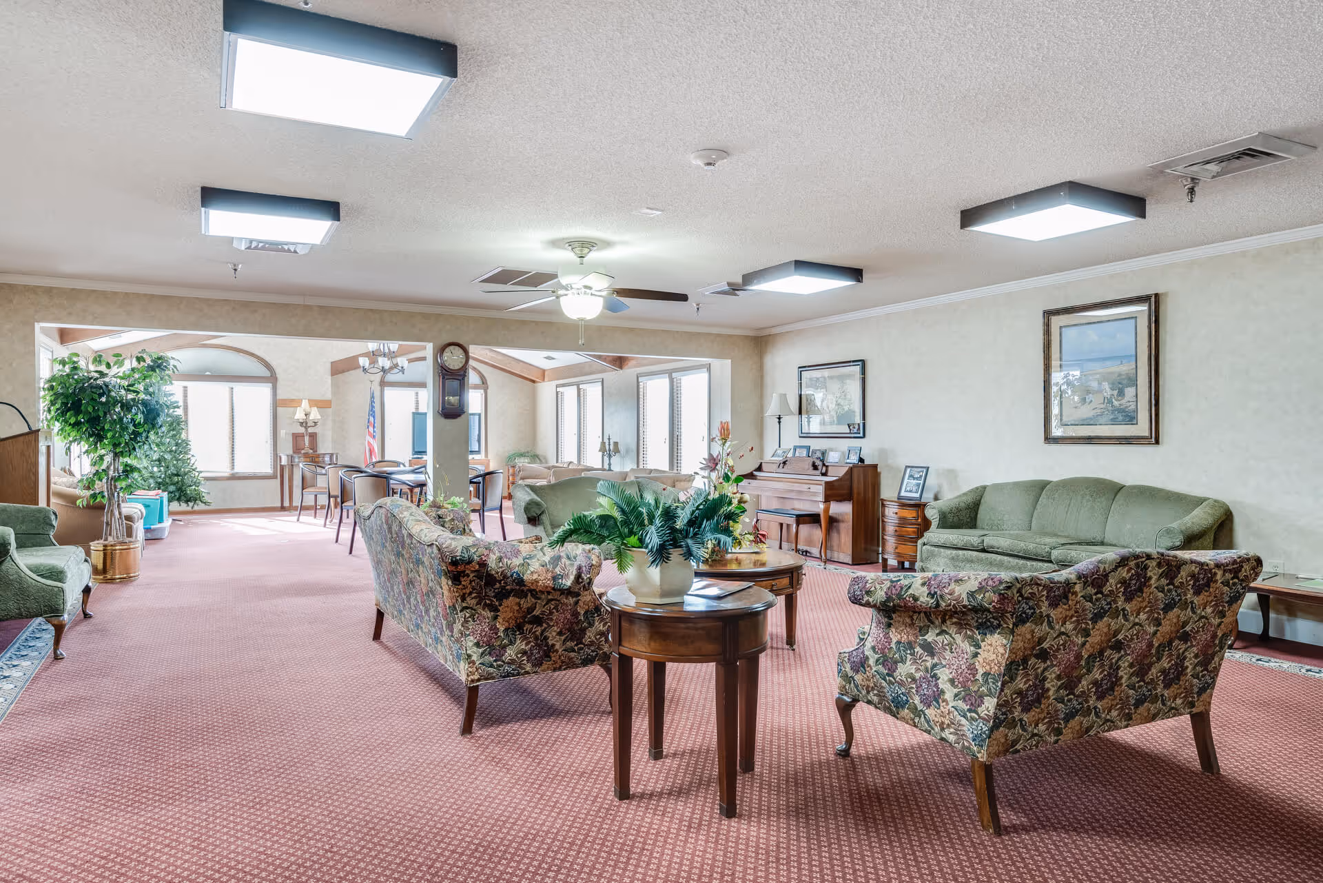 Spacious common lounge with floral upholstered sofas, a green couch, side tables, piano, plants, and large windows.