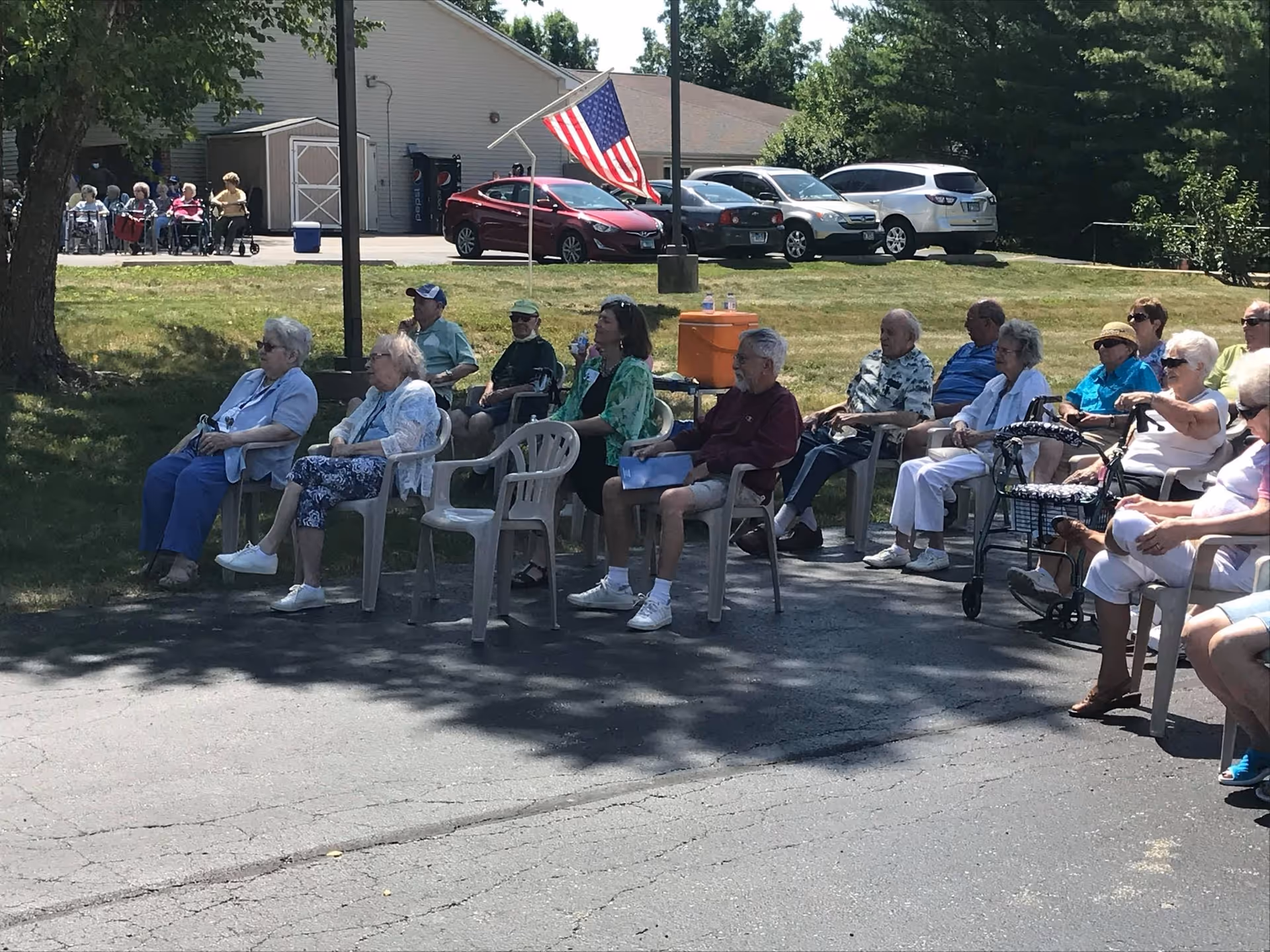 A group of elderly people sitting outdoors on plastic chairs and some in wheelchairs, gathered in a parking lot area near a building with parked cars and an American flag on a pole. The scene is sunny with trees and grass in the background.