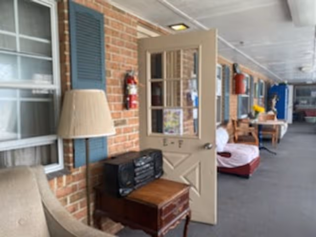 A covered outdoor hallway with brick walls and windows, featuring a beige door labeled 'E-F' that is open. There is a wooden side table with a stereo system and a lamp next to a beige armchair. Further down the hallway, there are chairs and tables with decorations, including a vase with yellow flowers.