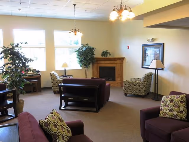 A cozy living room area in an assisted living facility featuring several upholstered chairs and sofas with patterned cushions, a wooden fireplace with a plant on top, large windows letting in natural light, potted plants, a piano in the corner, and ceiling light fixtures.