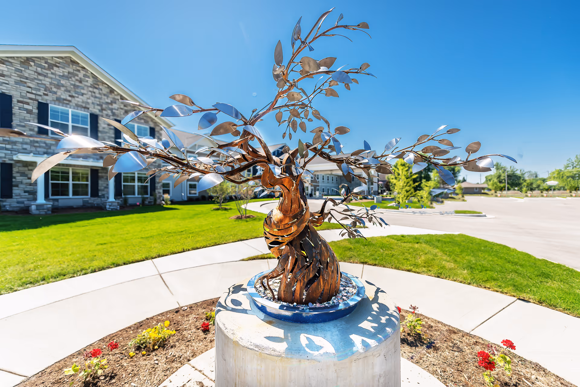A metal sculpture of a tree with twisting branches and leaves, placed on a circular concrete pedestal in a landscaped garden area outside a stone building under a clear blue sky.