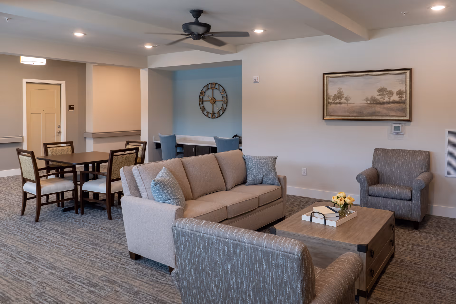 A cozy living room area with a beige sofa, two armchairs, and a wooden coffee table with books and a small flower arrangement. In the background, there is a dining table with four chairs, a wall clock, and a framed landscape painting on the wall. The room has neutral-colored walls and carpeted flooring.
