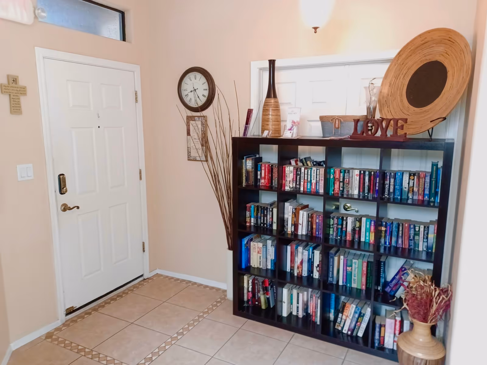 An interior entryway with a white door on the left and a black bookshelf filled with books on the right. Above the bookshelf are decorative items including a large round wicker tray, a vase, and a wooden sign spelling 'LOVE'. A clock and framed artwork hang on the wall above a bundle of tall decorative sticks in the corner. The floor is tiled with a patterned border.