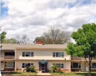 Exterior view of a two-story assisted living facility building with a beige facade, multiple windows, a central entrance with a green awning, surrounded by trees and greenery under a cloudy sky.