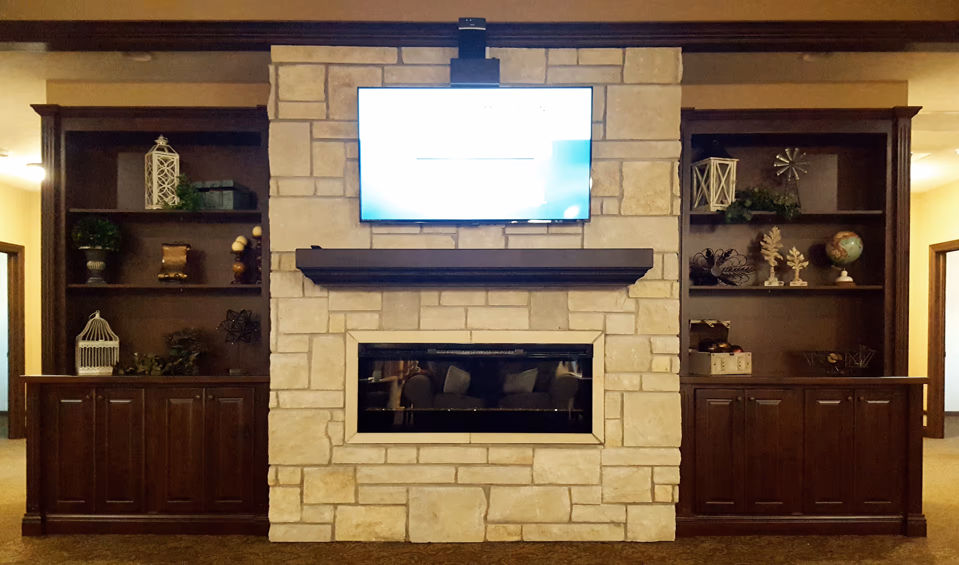 Stone fireplace with a mounted TV above flanked by dark wood built-in shelves and cabinets in a communal living area.