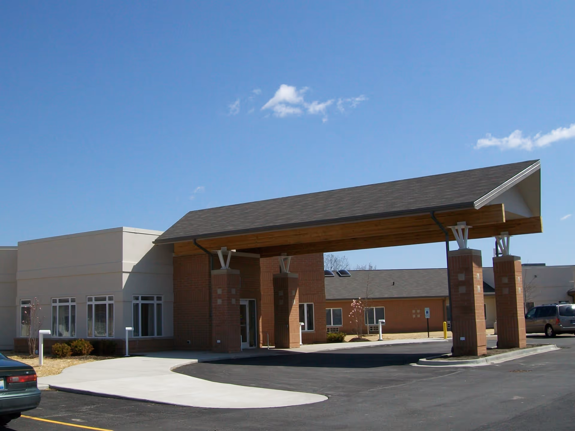 Exterior view of a senior living facility entrance with a large covered drop-off area supported by brick pillars, adjacent to a parking lot under a clear blue sky.