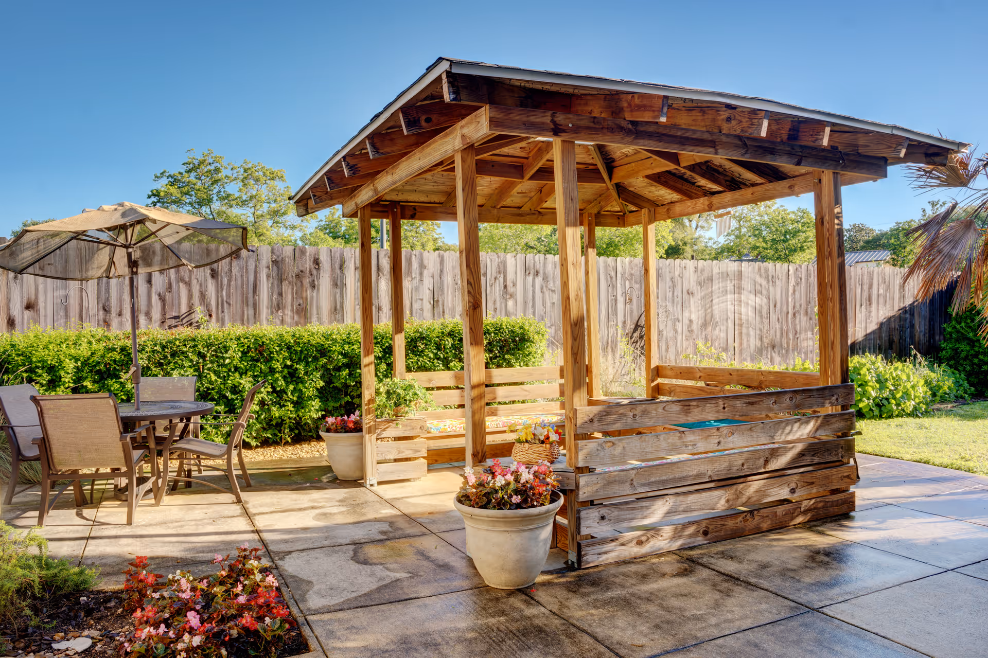 Outdoor patio area with a wooden gazebo featuring built-in bench seating, surrounded by potted plants and flowers. To the left, there is a round table with four chairs and a large umbrella. The area is enclosed by a wooden fence with green bushes along the fence line under a clear blue sky.