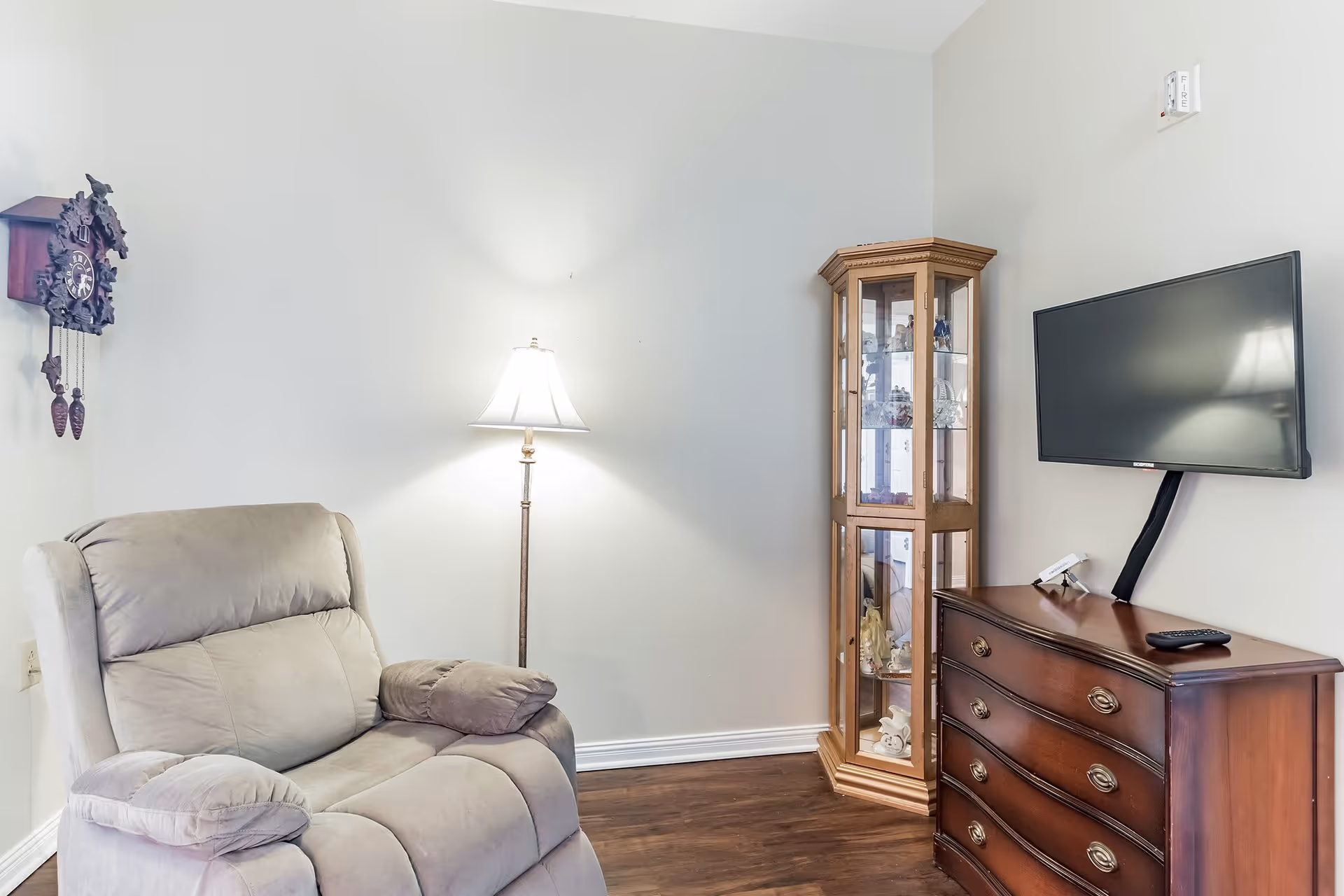 A cozy living room corner featuring a beige recliner chair, a floor lamp with a white shade, a wooden display cabinet with glass doors showcasing decorative items, a wall-mounted flat-screen TV, and a wooden dresser with three drawers and a remote control on top. A cuckoo clock hangs on the wall to the left.
