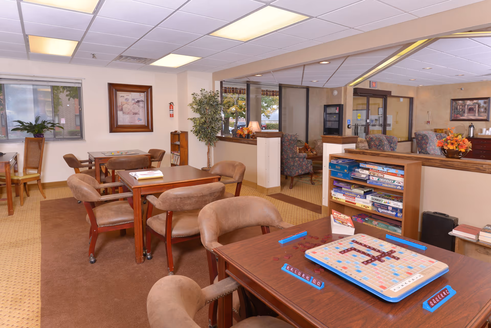 A cozy senior living common area with multiple tables and chairs arranged for playing board games like Scrabble. There are shelves filled with various board games, comfortable armchairs, a window with a view outside, and decorative plants and artwork on the walls. The room has a warm, inviting atmosphere with soft lighting from ceiling fixtures.