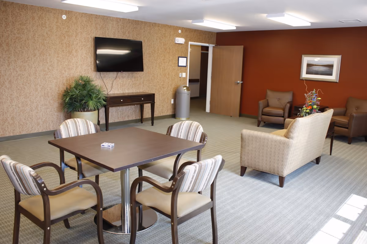 A senior living facility common room with a square table surrounded by four striped cushioned chairs in the foreground. In the background, there is a wall-mounted flat screen TV above a small dark wooden table, a large potted plant, a trash can, and an open door. To the right, there are three armchairs arranged around a small table with a flower arrangement, and a framed picture on a red accent wall.