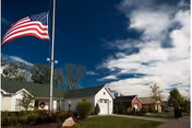 American flag flying beside single-story residential buildings and a walkway under a partly cloudy sky.