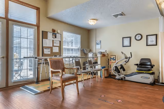 A rehabilitation or therapy room with wooden flooring, featuring a stationary exercise bike, parallel bars for walking support, several chairs, a bulletin board with notices, and large windows letting in natural light.