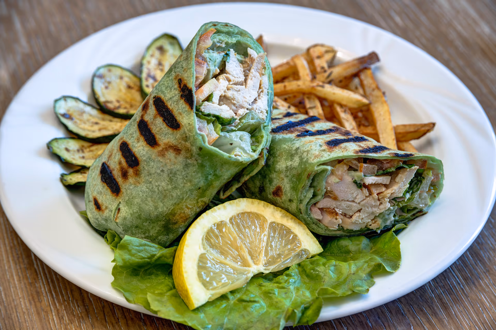 A white plate with a grilled chicken wrap cut in half, served with grilled zucchini slices, French fries, a lemon wedge, and a lettuce leaf on a wooden table.