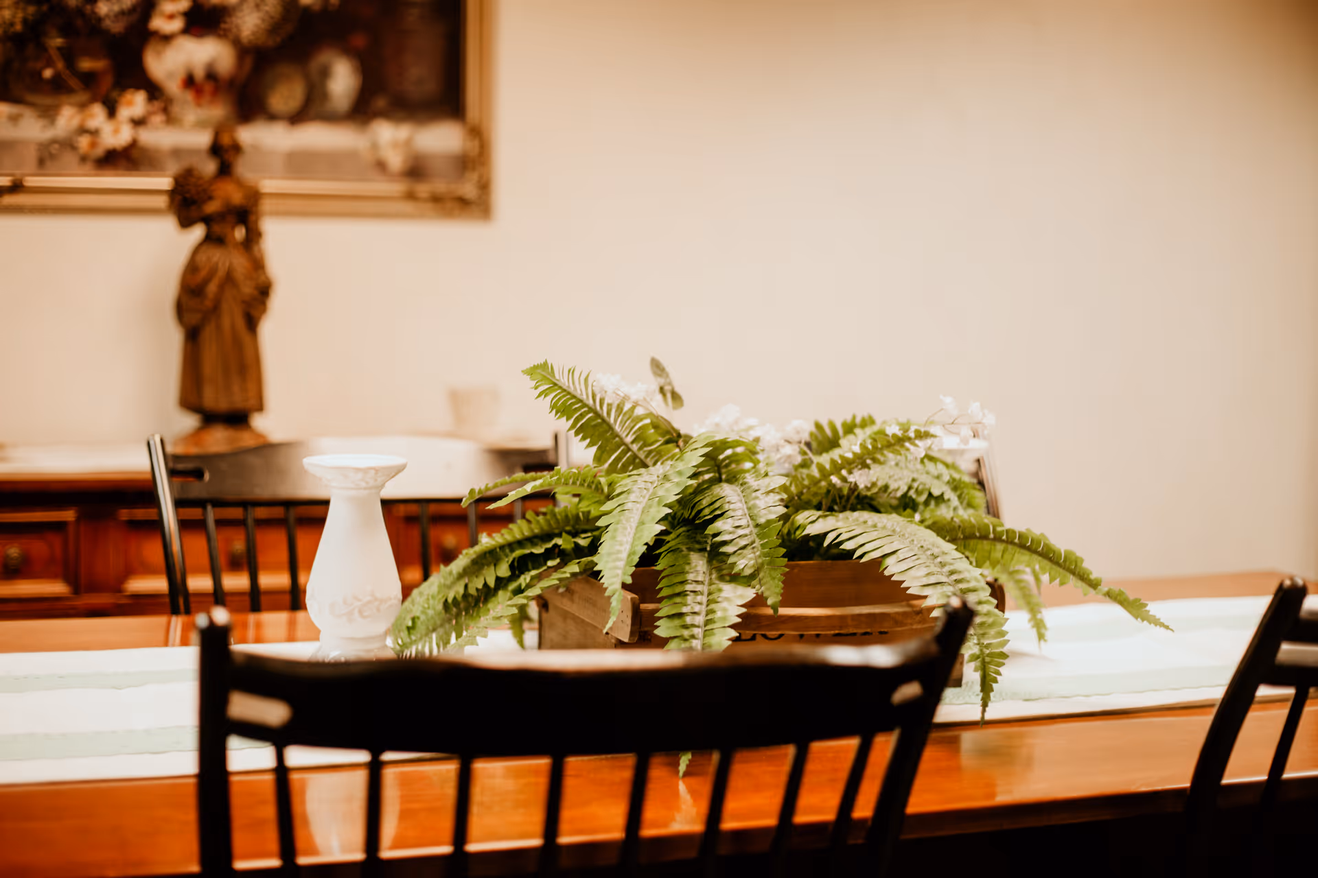 A wooden dining table with a green and white table runner, decorated with a wooden planter box containing green ferns and white flowers. Black wooden chairs surround the table. In the background, there is a wooden sideboard with a decorative statue and a framed painting on the wall.