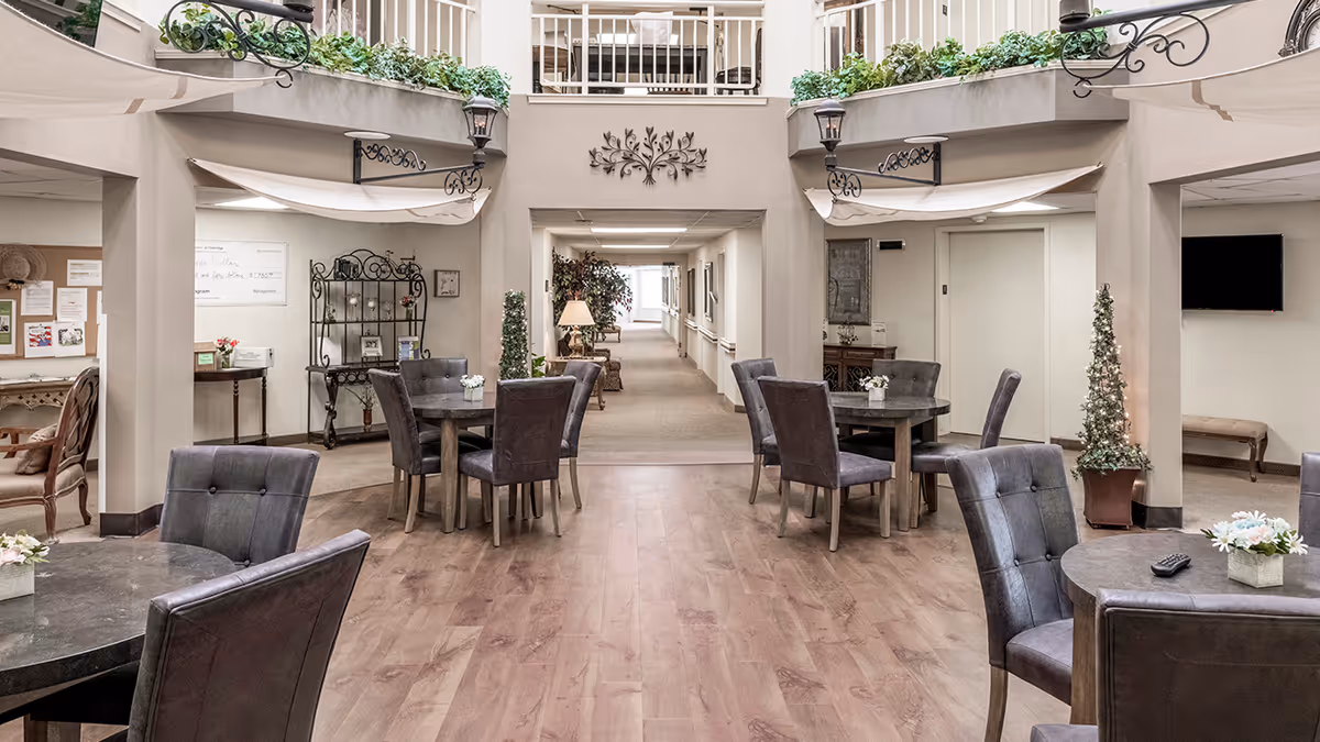 Interior common area of a senior living facility with round tables and chairs arranged on a wooden floor. The space features decorative plants, wall art, and a hallway leading to other rooms. There is a balcony with railings and greenery above, and soft lighting throughout the area.