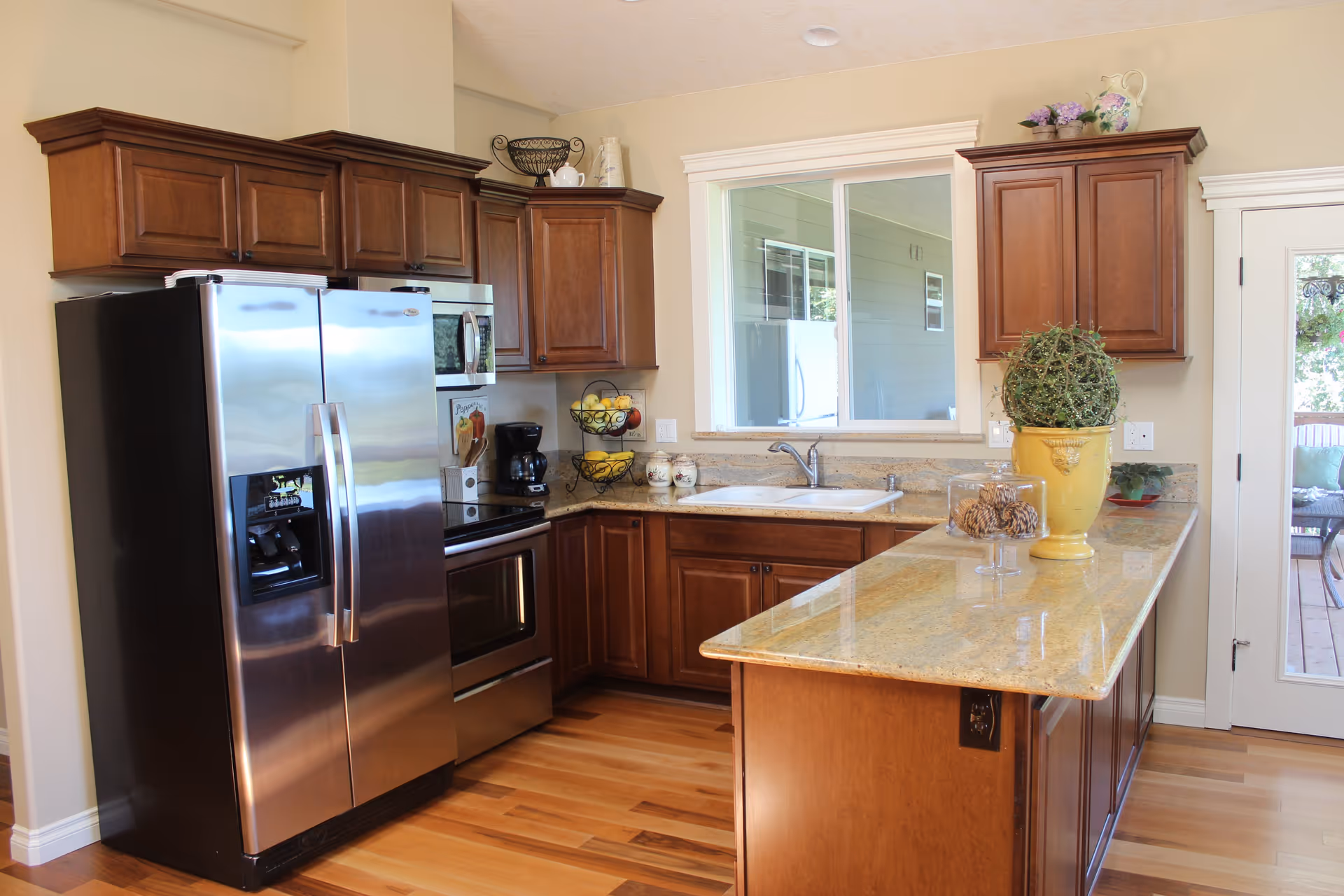 A modern kitchen with wooden cabinets, a stainless steel refrigerator, oven, and microwave. There is a granite countertop with a yellow potted plant and a glass container with pine cones. A window above the sink looks out to an outdoor area, and a glass door leads to a patio with seating.