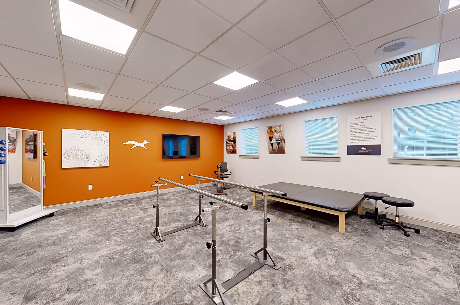 A physical therapy or rehabilitation room with parallel bars for walking exercises, a padded therapy table, two stools, an exercise bike, and a large mirror on wheels. The room has a bright orange accent wall with a mounted TV and a fox logo, and the other walls are white with windows and motivational posters.