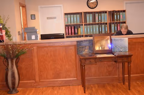 Reception area with a wooden front desk, a person working behind the desk, a small wooden table with framed pictures on it, a large vase with decorative branches on the left, and shelves filled with binders and a clock on the wall behind the desk.