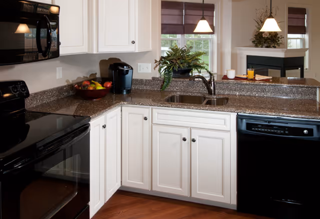 Modern kitchen with white cabinets, granite countertops, a black stove, microwave, dishwasher, a double sink, a coffee maker, and a bowl of fruit. In the background, there are windows with brown blinds and a dining area with pendant lights and a decorative plant.