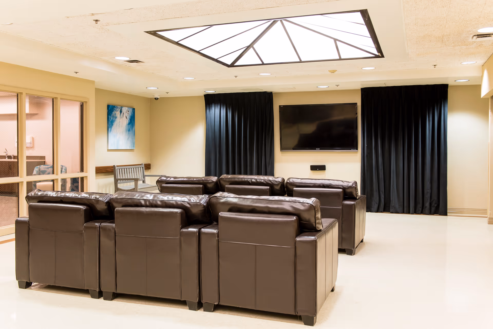 A lounge with rows of brown leather armchairs facing a wall-mounted TV under a skylight, with black curtains and pale walls.