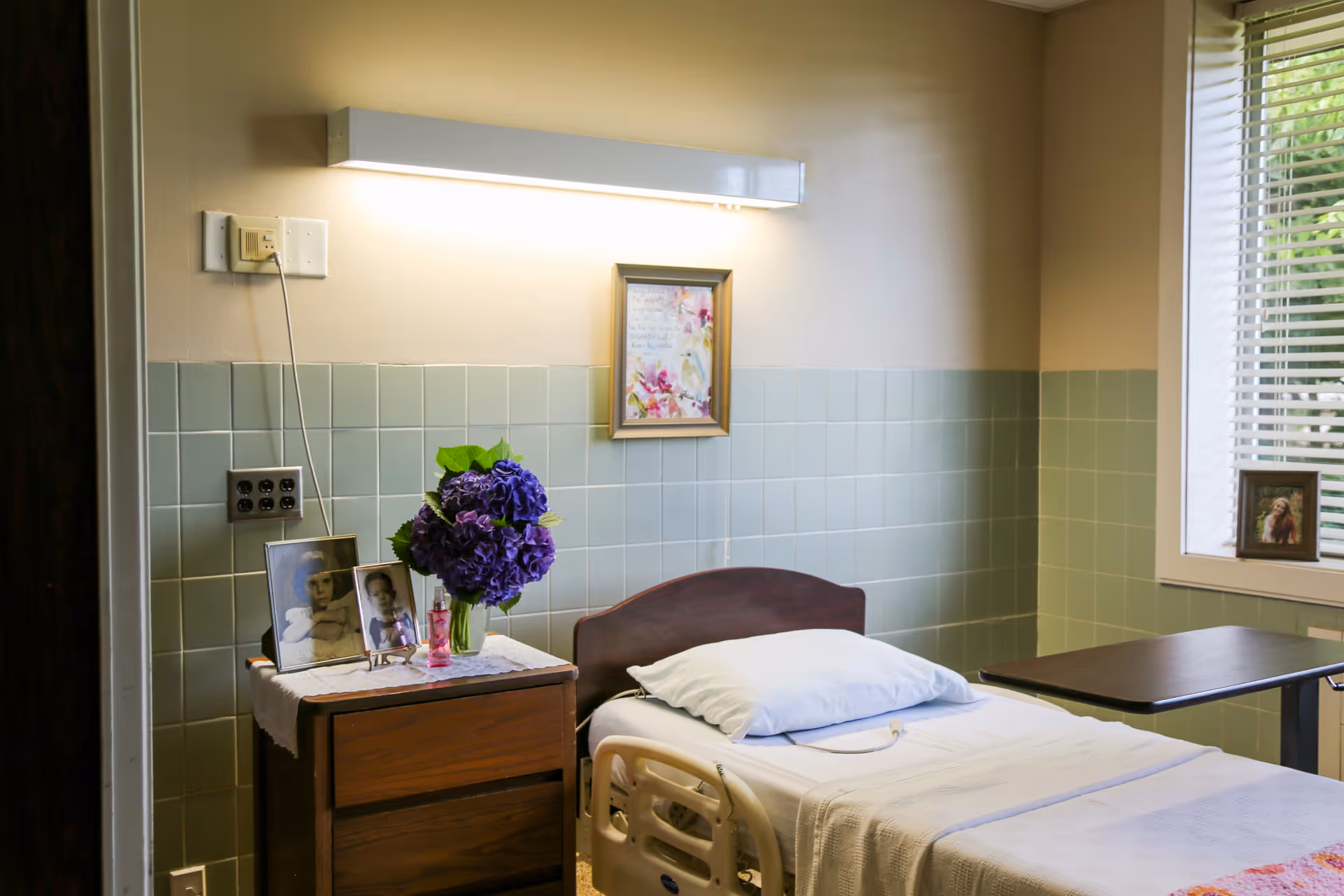 A tidy senior care bedroom with a single hospital-style bed, bedside table holding framed photos and a vase of purple flowers, and a window with blinds.