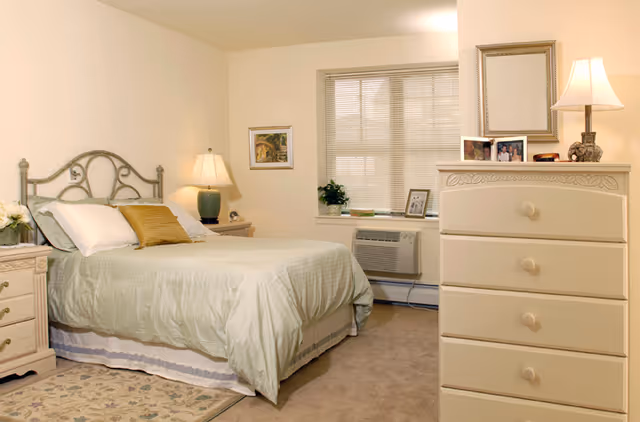 A neatly made bed with light green bedding and a mustard yellow pillow in a bedroom. There is a nightstand with a lamp and flowers on the left side of the bed. A window with blinds and a small plant on the windowsill is visible behind the bed. On the right side, there is a cream-colored dresser with a lamp, picture frames, and a mirror above it. The room has beige carpet and light-colored walls.