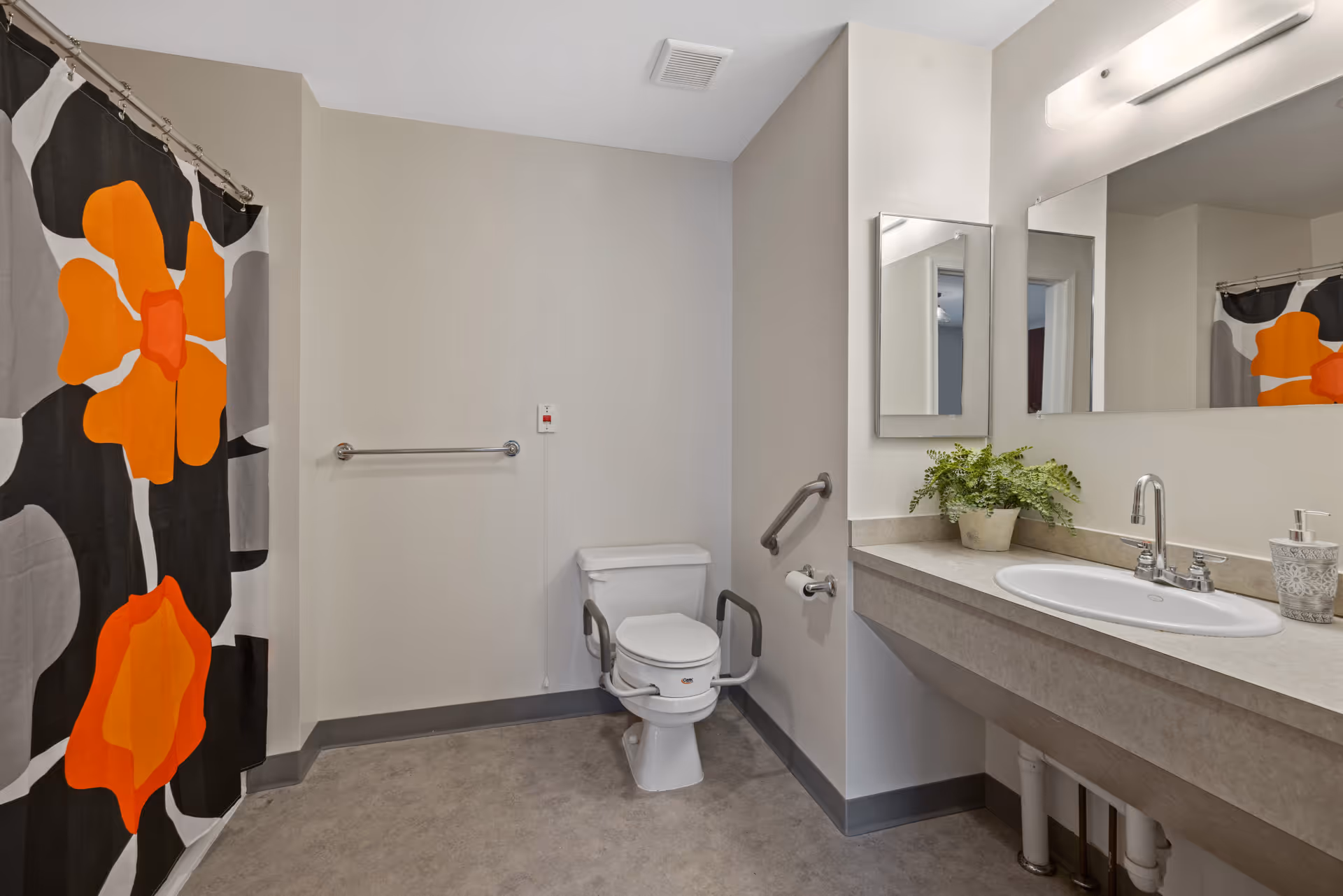 A clean and accessible bathroom featuring a toilet with support rails, a sink with a large mirror above it, a soap dispenser, a small potted plant on the countertop, and a shower with a curtain decorated with large orange and black floral patterns.