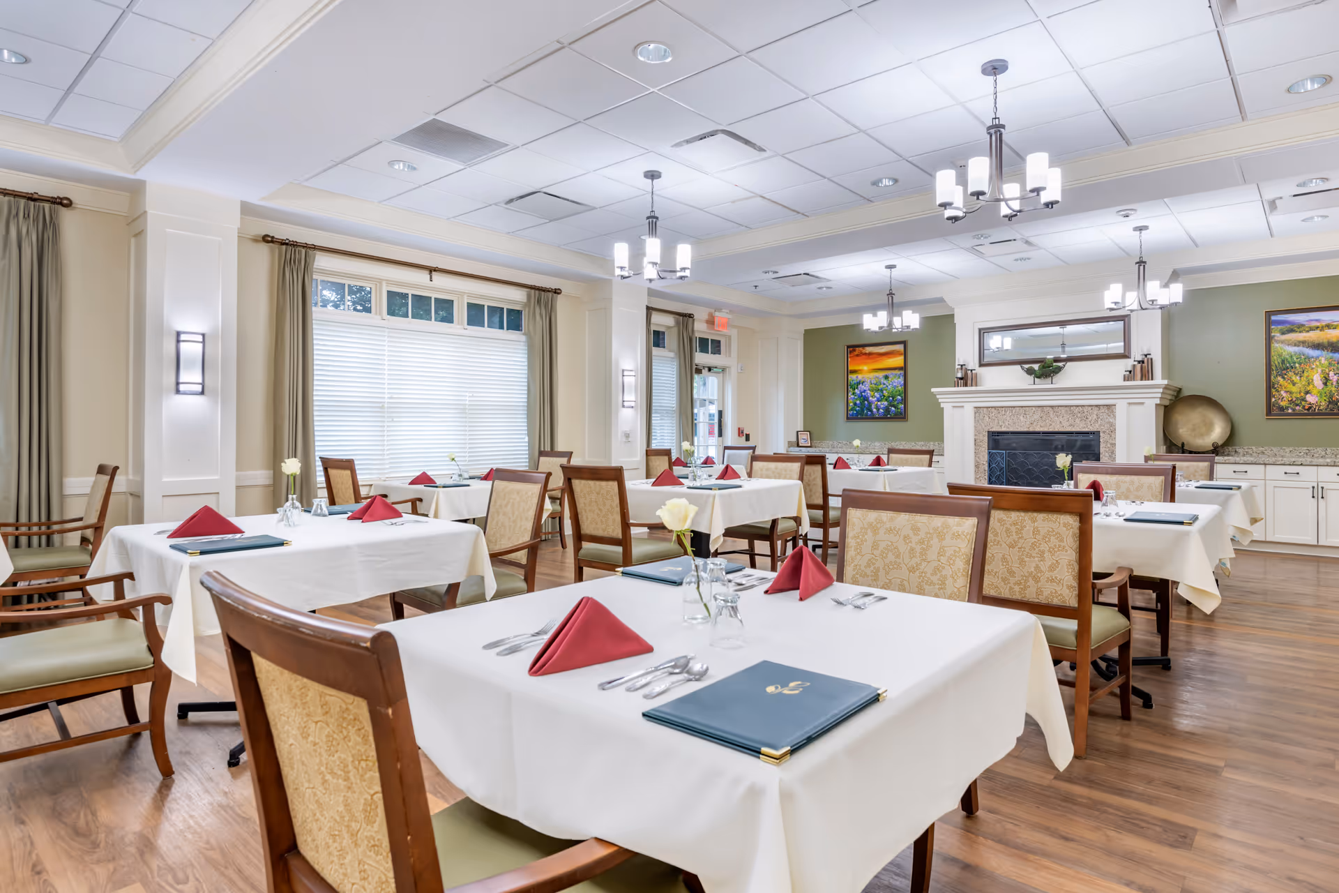 Bright dining room with white-tableclothed tables set with red napkins and wooden chairs in a senior living facility.