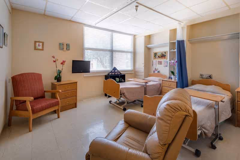A nursing facility room with two hospital-style beds, each with a small overbed table. The room has a large window with blinds, a red armchair, a beige recliner, two wooden dressers, and some decorative items including flowers and framed pictures on the walls.