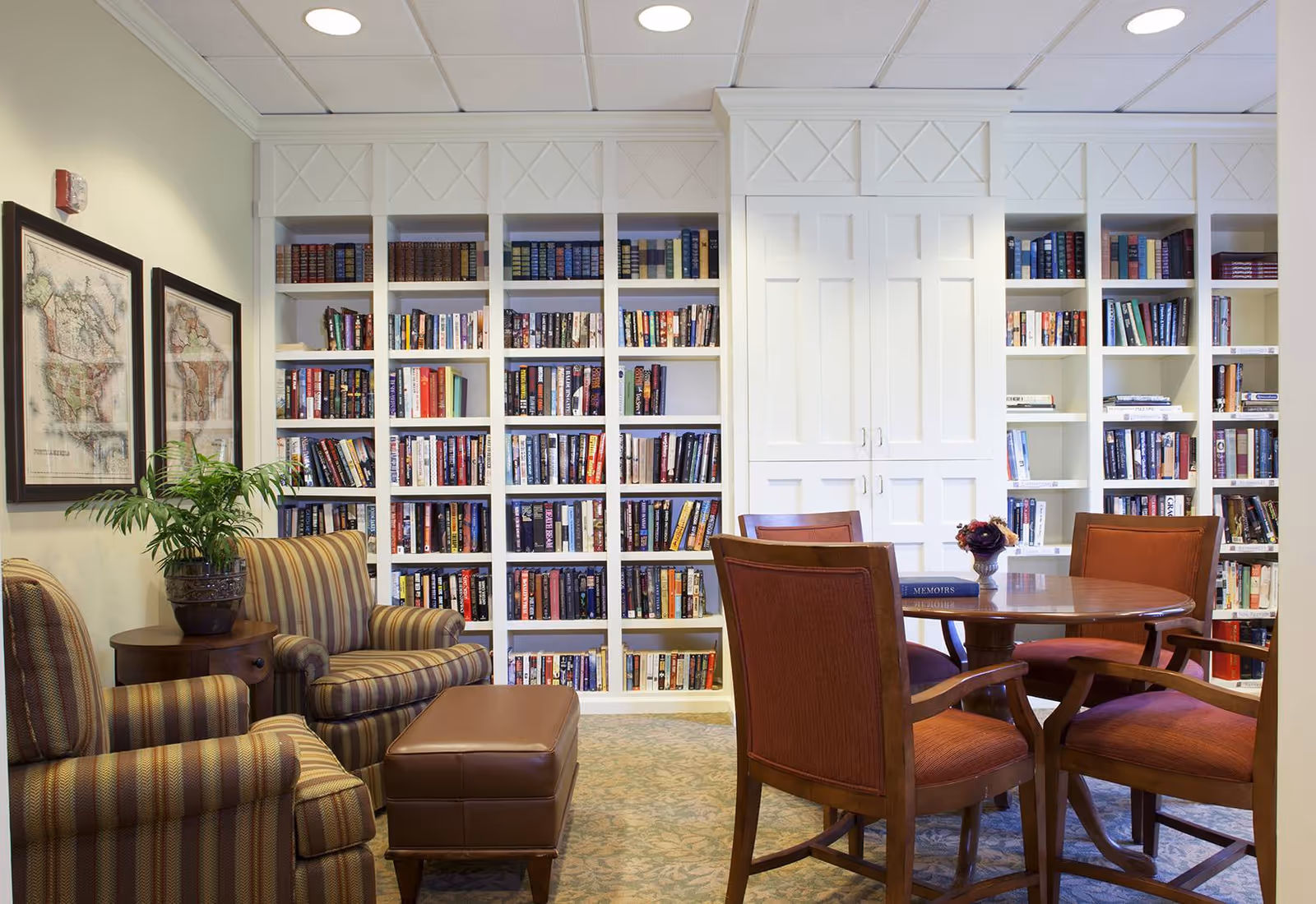 Cozy reading room with floor-to-ceiling bookshelves, a round table with four chairs, and striped armchairs with an ottoman.